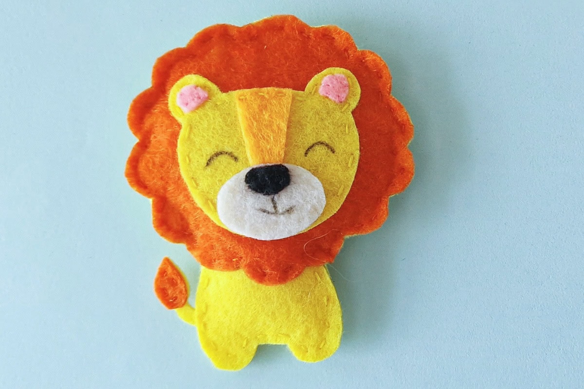 Finished yellow felt lion plush with an orange mane and tufted tail on a blue background
